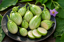 Load image into Gallery viewer, A plate of fresh striped eggplant vegetables, including whole elongated eggplants and sliced rounds, arranged on a dark ceramic plate with green leaves and a purple flower around it.